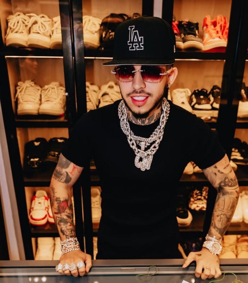 Westcol con gorra negra, gafas rosadas y cadenas, frente a una vitrina de tenis